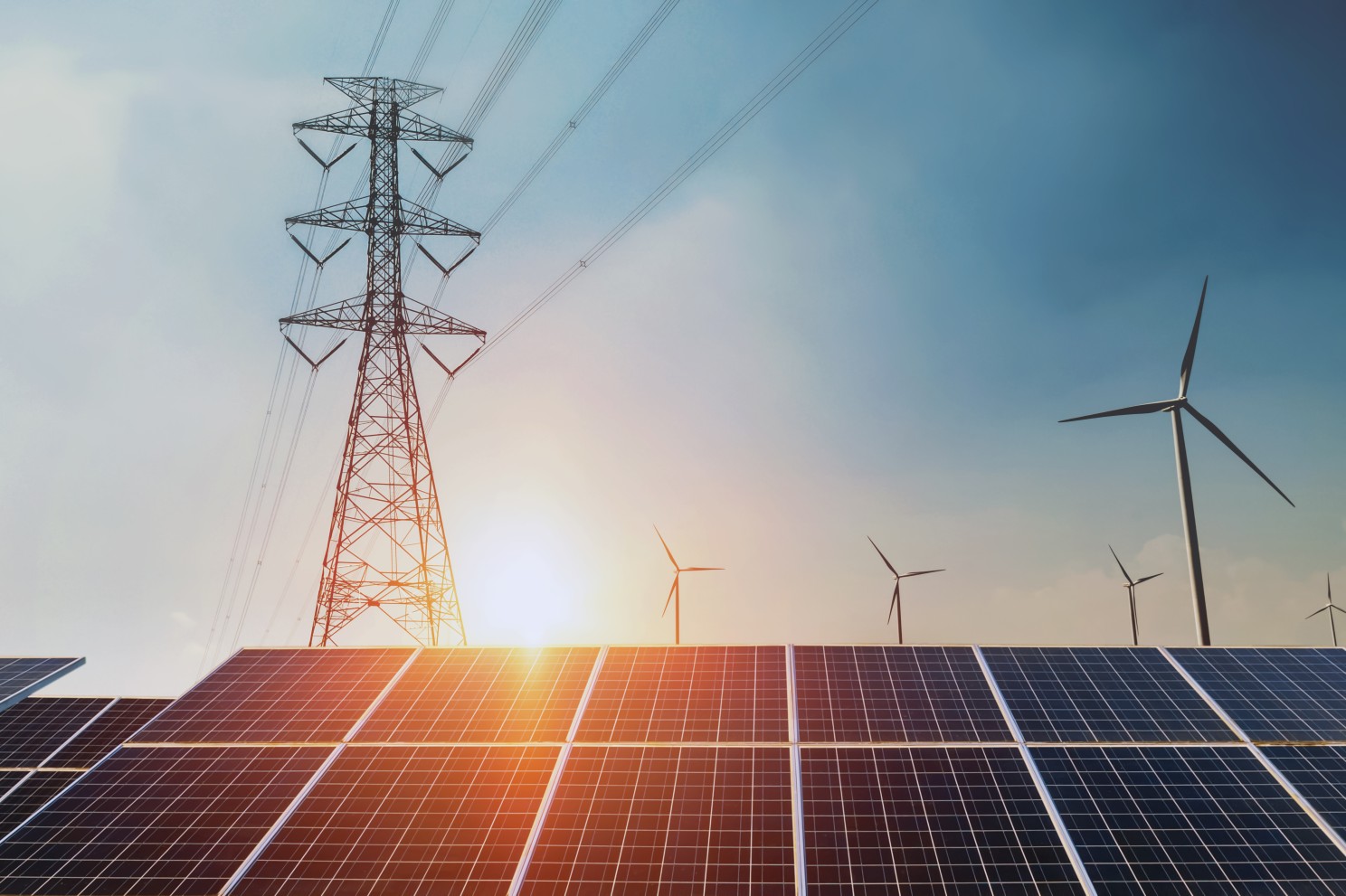 Renewable energy landscape featuring solar panels in the foreground, wind turbines in the background, and a high-voltage power transmission tower under a clear blue sky during sunset.